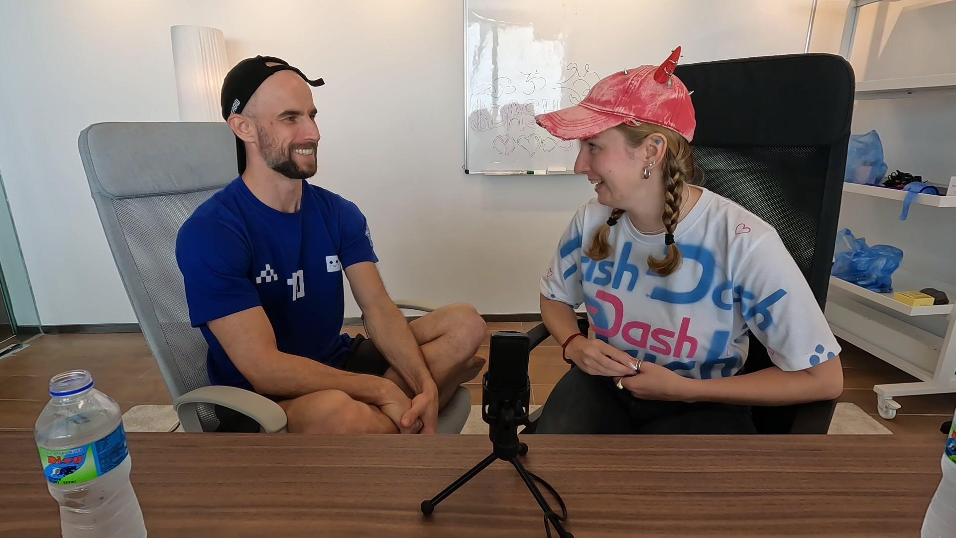 Daria Chernozub, in a Dash t-shirt and pink cap, laughing with Konrad during the podcast, a whiteboard reading $DASH behind them